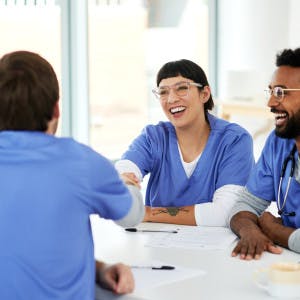 A photograph of a diverse group of nurses around a table, where a man and woman sat opposite each other are shaking hands as to welcome a new hire